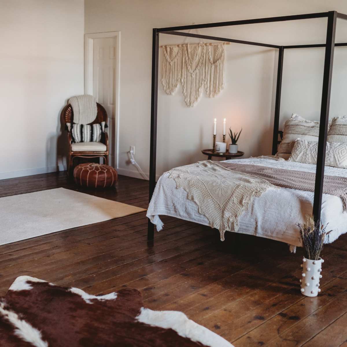 A minimalistic bedroom featuring a four-poster bed, a woven wall hanging, a chair, and a floor rug on wooden flooring.