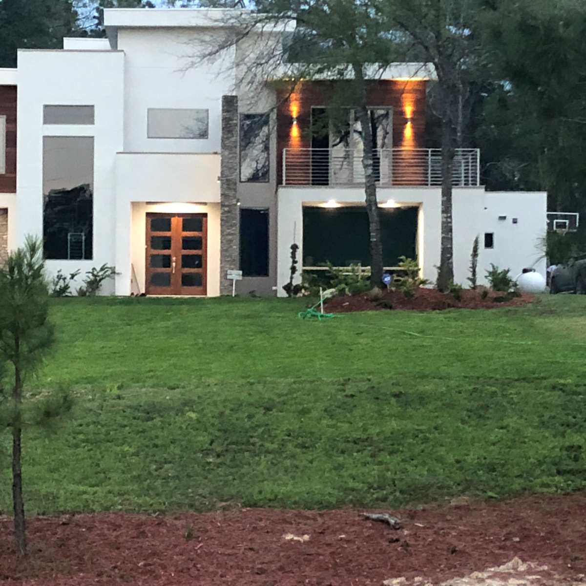A modern two-story house features a combination of white walls, wooden accents, and stone details, with landscaped greenery in the front yard.