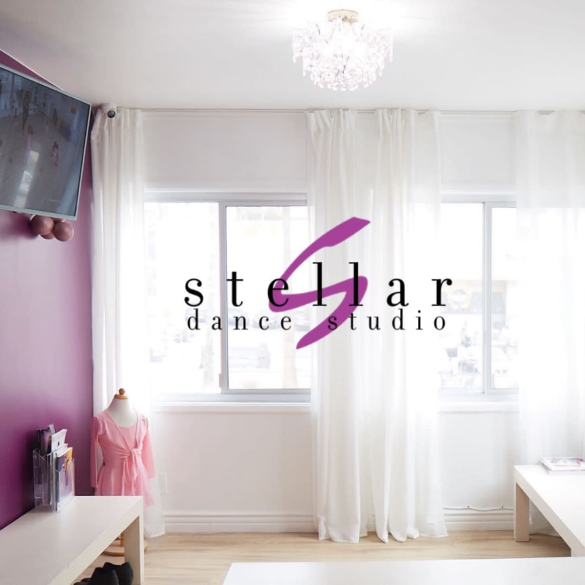 A dance studio with a purple accent wall, large windows covered with white curtains, and a small dress form wearing a pink outfit.
