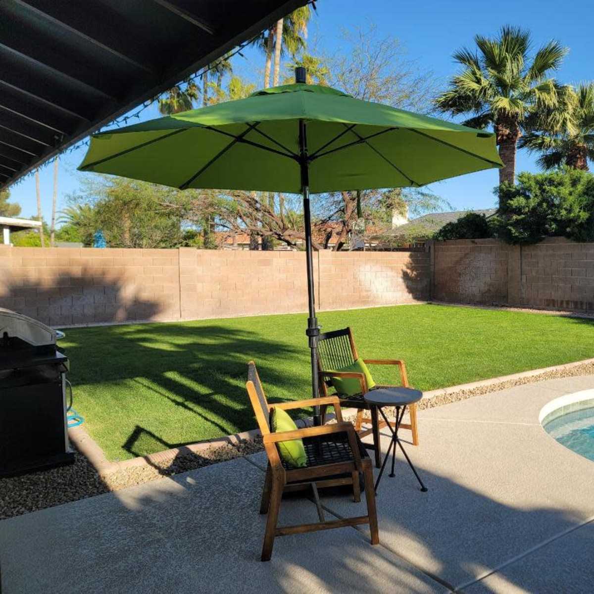 A green umbrella shades a small seating area with two chairs next to a swimming pool and a lawn surrounded by a brick wall and palm trees.