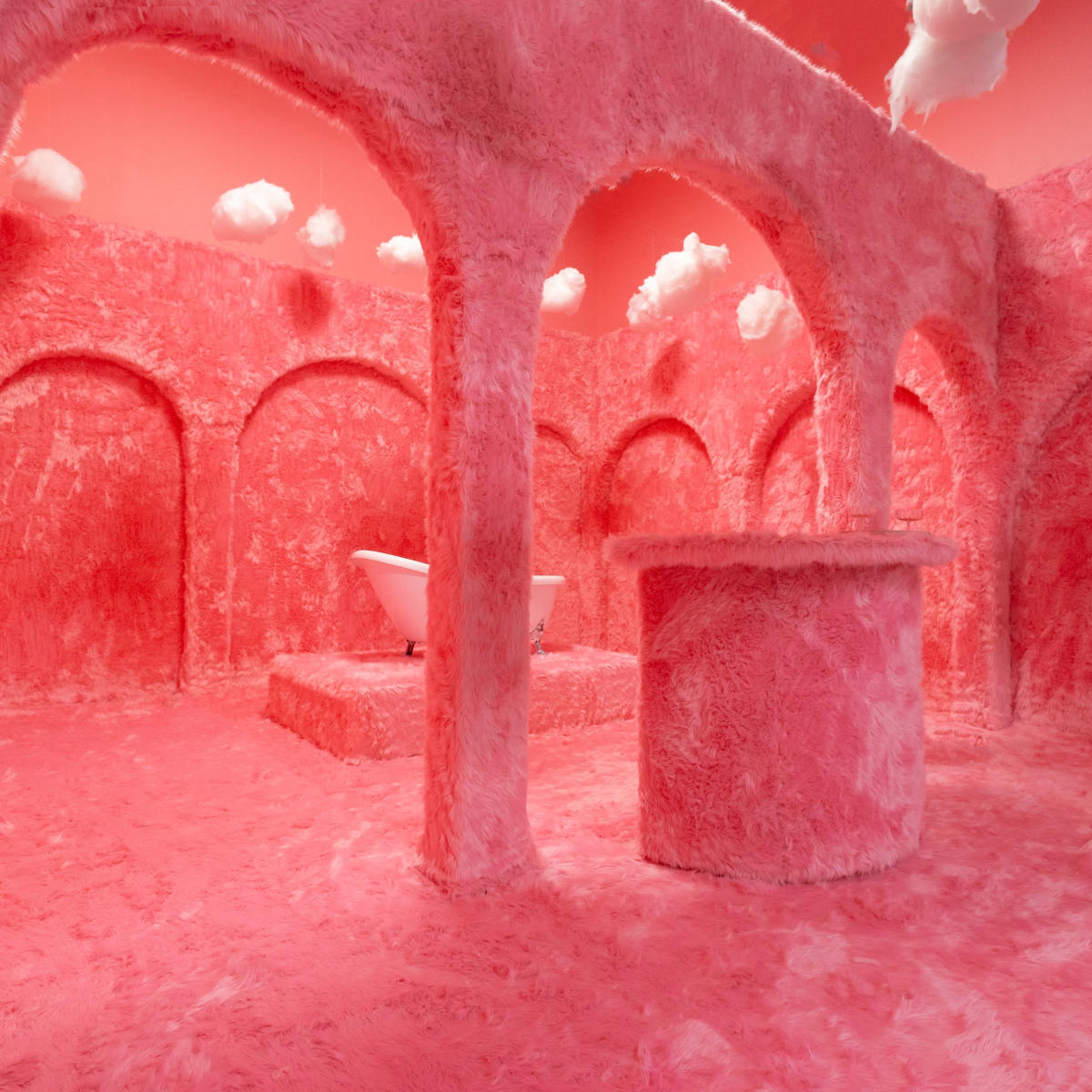 A surreal pink room with textured walls and arches, featuring a bathtub on a raised platform and fluffy clouds suspended from the ceiling.