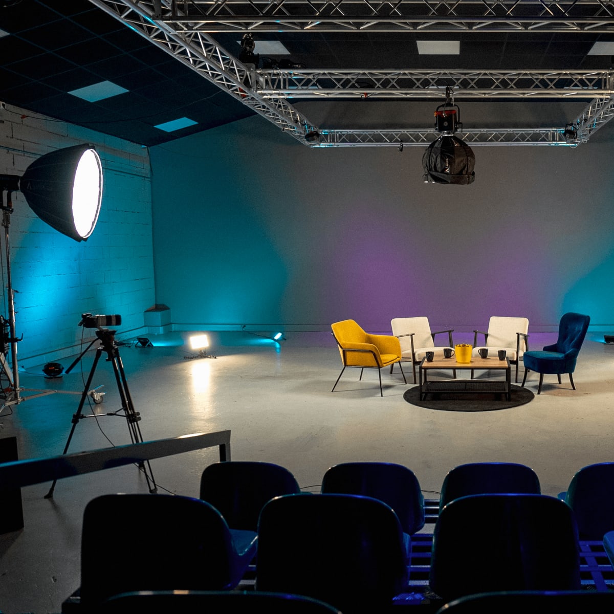 A modern studio setup features a circular seating arrangement with colored chairs, flanked by lighting equipment and cameras, against a neutral backdrop.