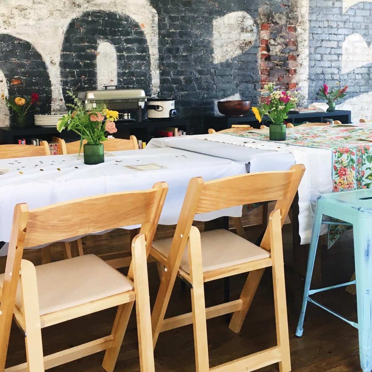 A rustic dining area with wooden folding chairs arranged around tables covered in white and patterned cloths, and small flower vases on each table.