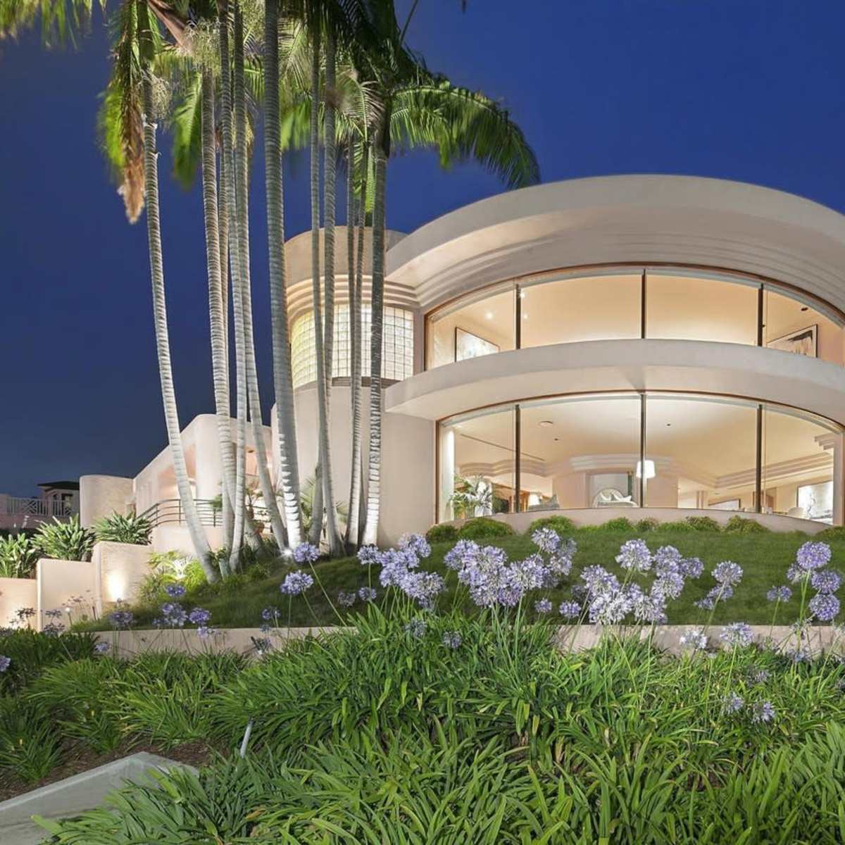A modern, circular house with large windows, surrounded by manicured gardens and palm trees at dusk.