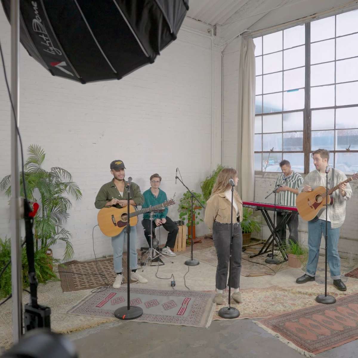 A group of musicians is performing with guitars and keyboards in a studio space featuring large windows and potted plants.