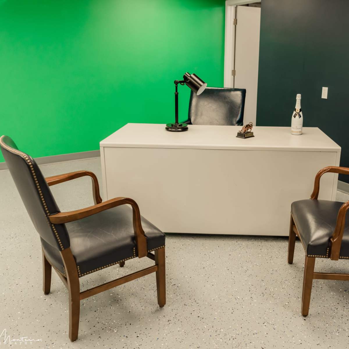 A small office space featuring a white desk, two wooden chairs, and a champagne bottle, with a green screen wall in the background.