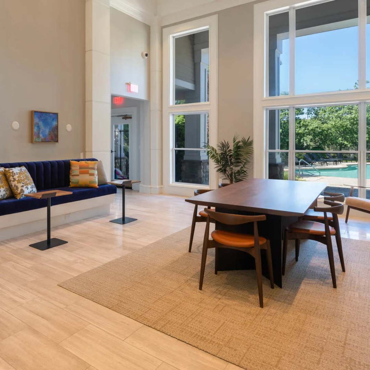 A modern lounge area featuring a blue upholstered bench, a wooden dining table with chairs, and large windows that overlook greenery.