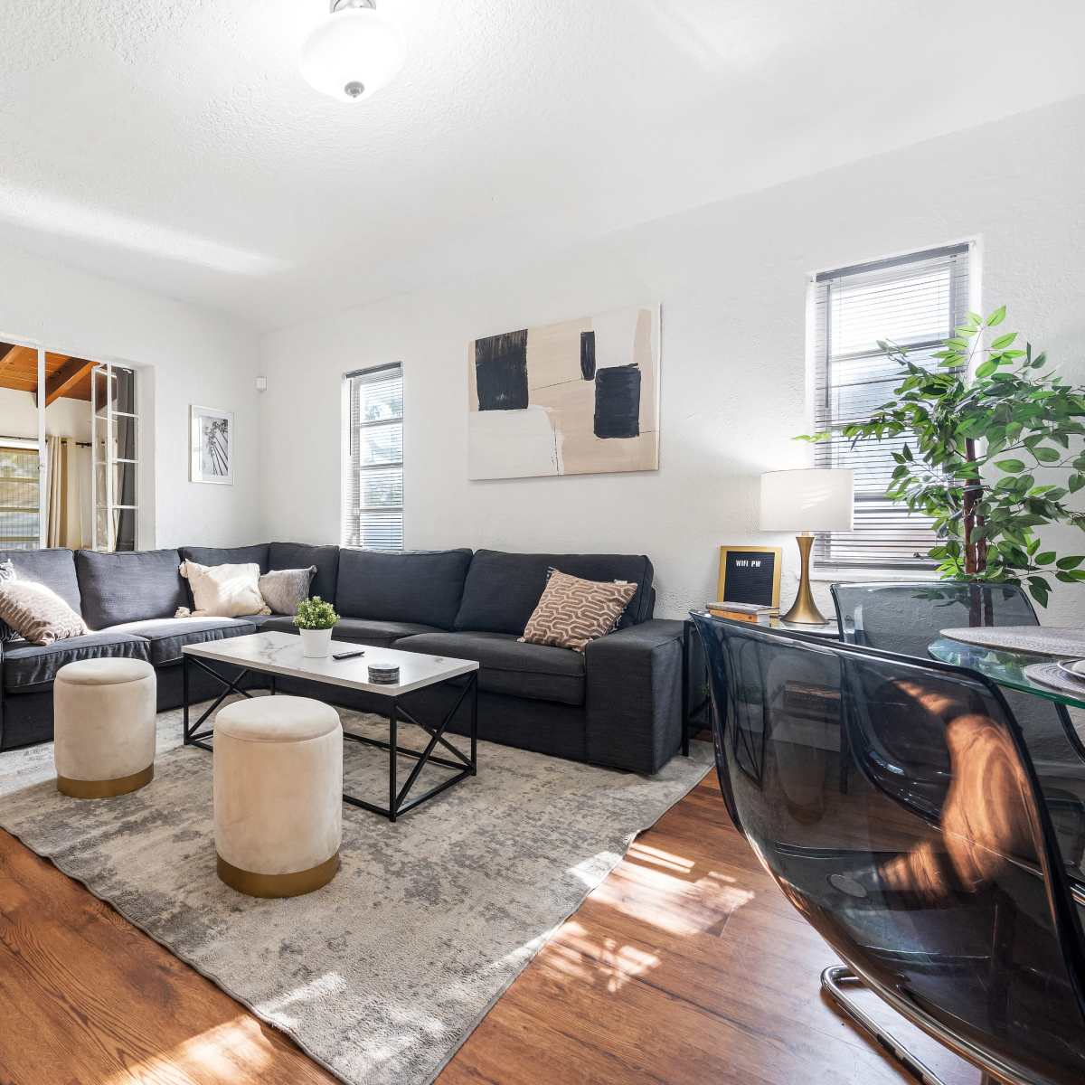 A modern living room features a dark sectional sofa, a coffee table, two ottomans, and a small desk, all set against white walls and hardwood flooring.