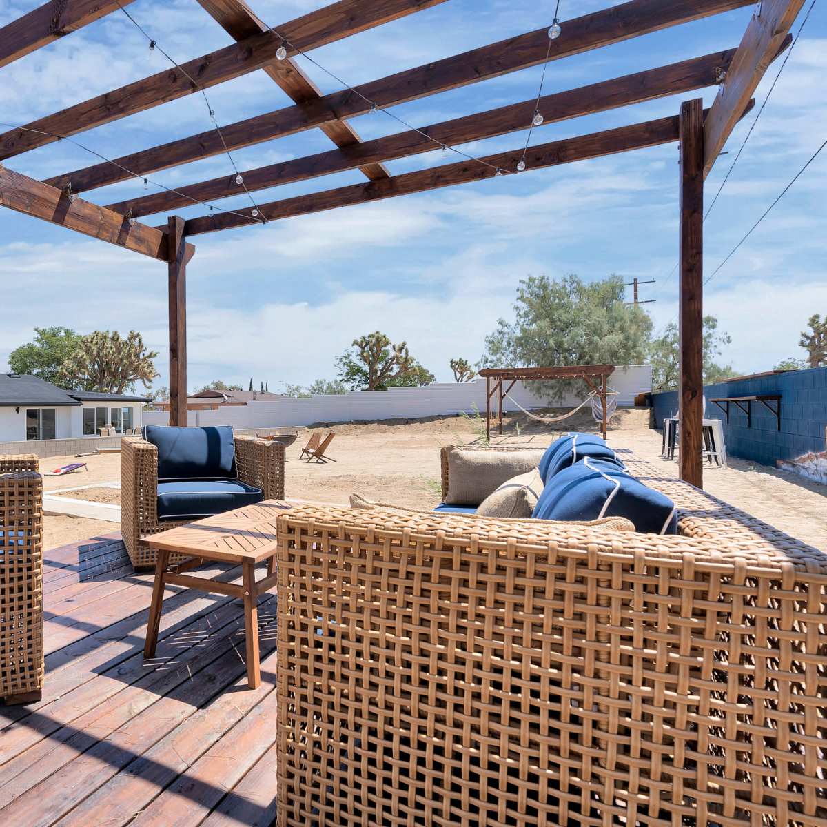 A wooden pergola with string lights frames a seating area featuring wicker chairs on a deck overlooking a barren backyard.