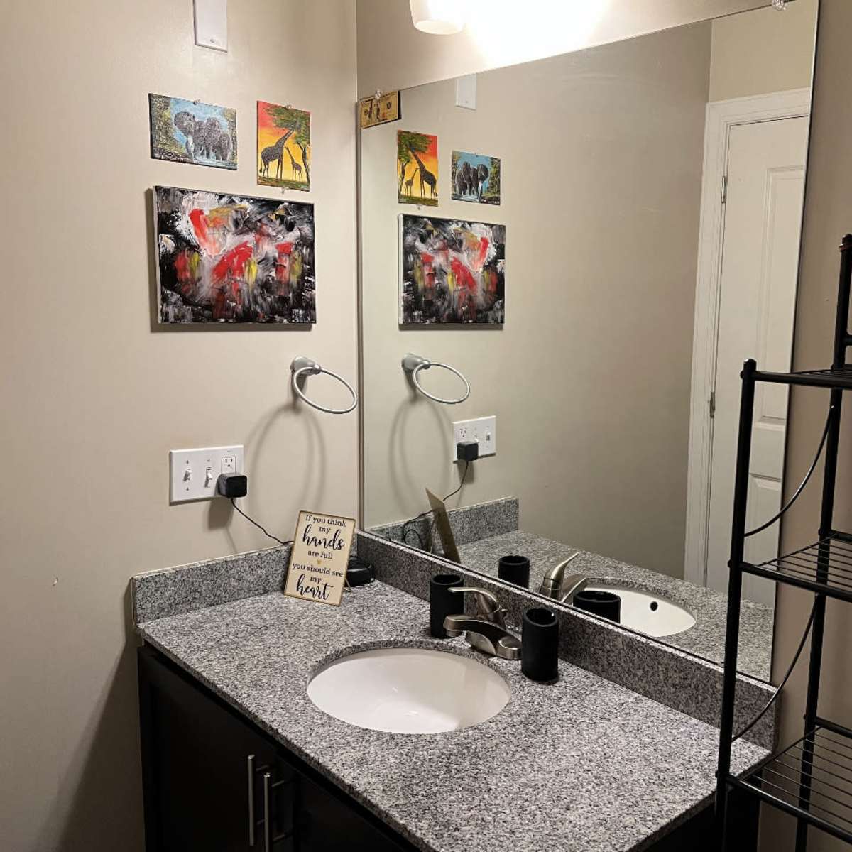 A bathroom features a granite countertop with a sink, a large mirror, two wall-mounted light fixtures, and artwork on the walls above.