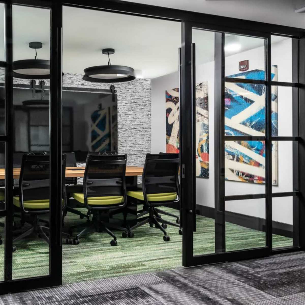 A glass-walled conference room features a wooden table surrounded by six black chairs with green accents, set against a backdrop of abstract artwork and a television screen.