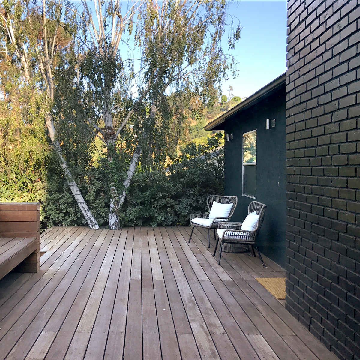 A wooden deck with a long bench, two chairs, and a backdrop of greenery and a tree.