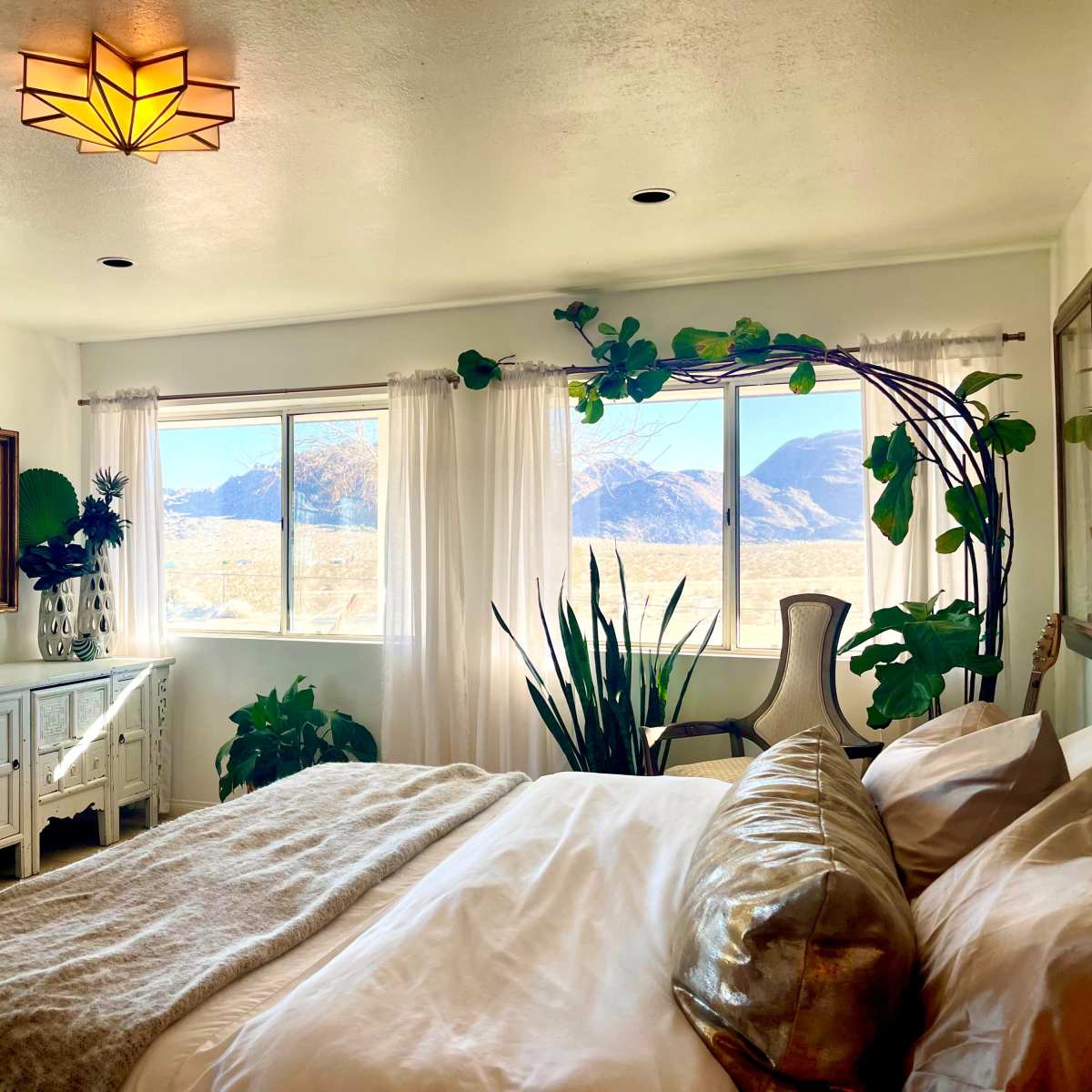 A bright bedroom features large windows with a view of mountains, adorned with indoor plants and a neatly made bed.