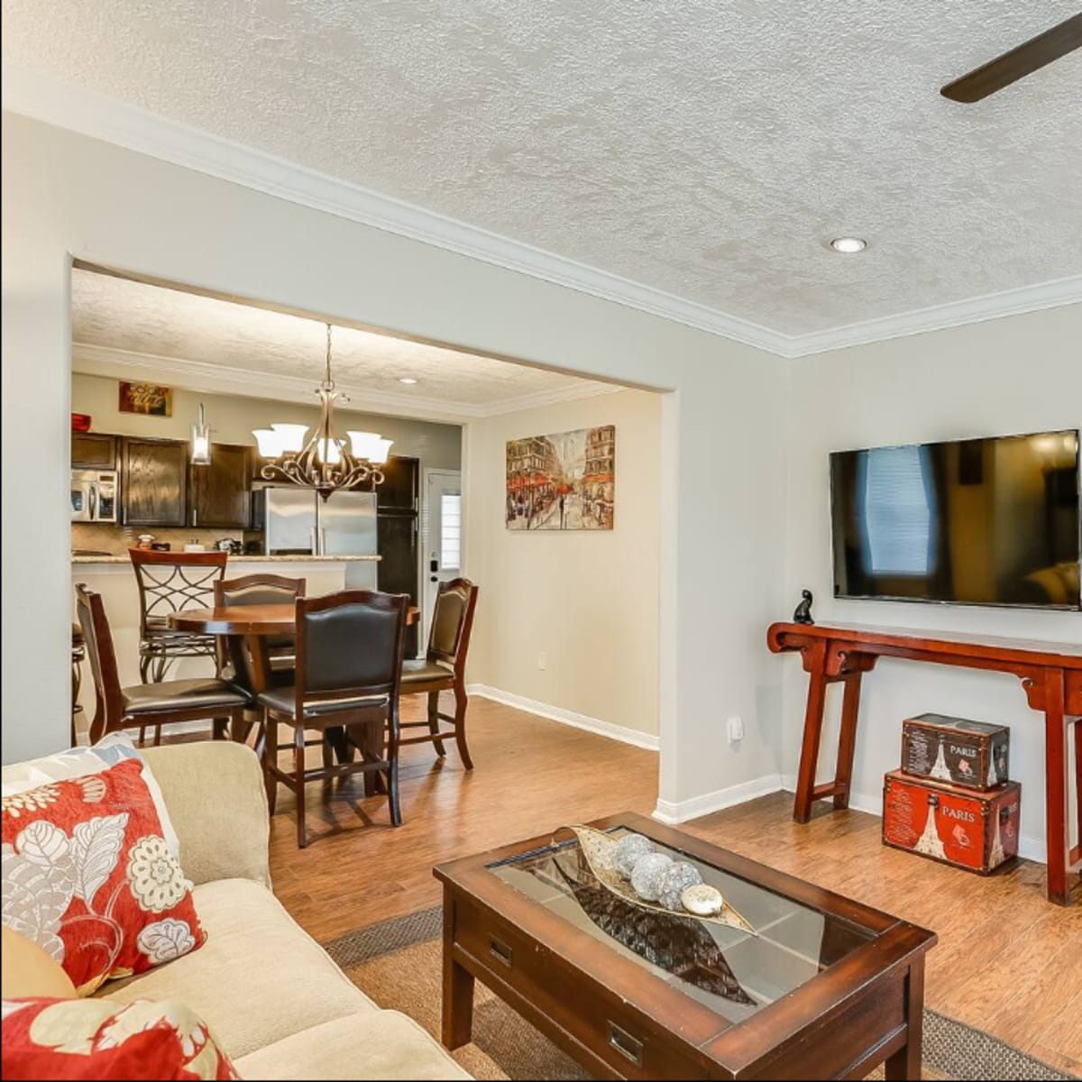 The living room features a beige sofa with red accents, a wooden coffee table, and a mounted TV on the wall, leading into a dining area and visible bedroom.