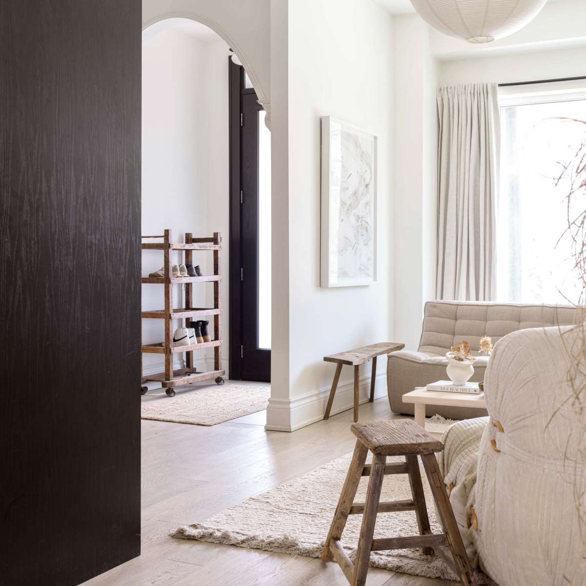A modern living room with a neutral color palette, featuring a sofa, a small wooden table, and a glimpse of a hallway with a wooden shelving unit.