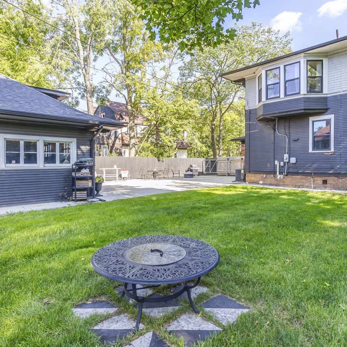 A backyard featuring a circular metal table on a stone patio, surrounded by a green lawn and a two-story house with a deck and a separate garage.