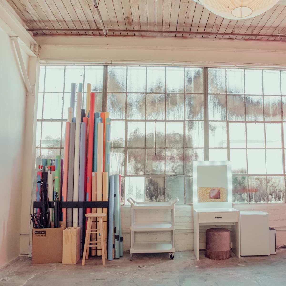 A workspace with a collection of colorful paper rolls stacked against a large, dusty window, alongside a desk area with a computer and chair.
