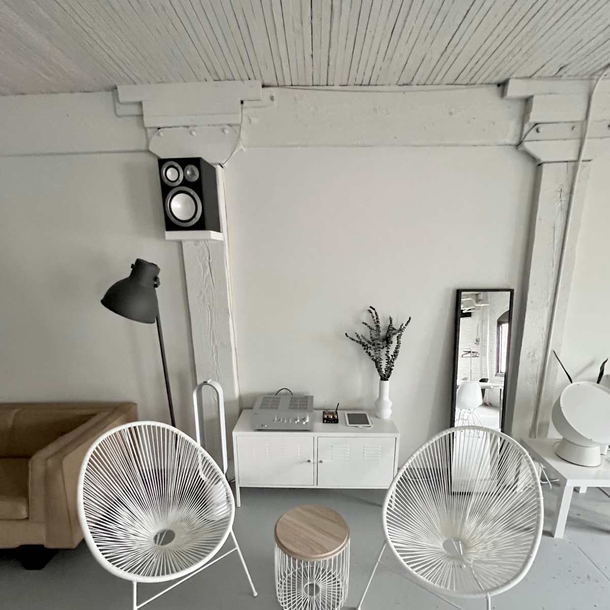 A minimalistic interior featuring two white chairs, a small wooden table, a floor lamp, a wall-mounted speaker, and a console table with a mirror and decorative plant.