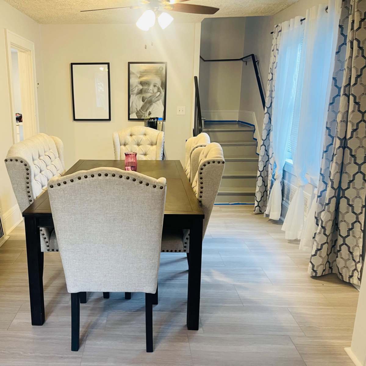 A dining room with a rectangular table surrounded by four upholstered chairs, a ceiling fan above, and a staircase visible in the background.