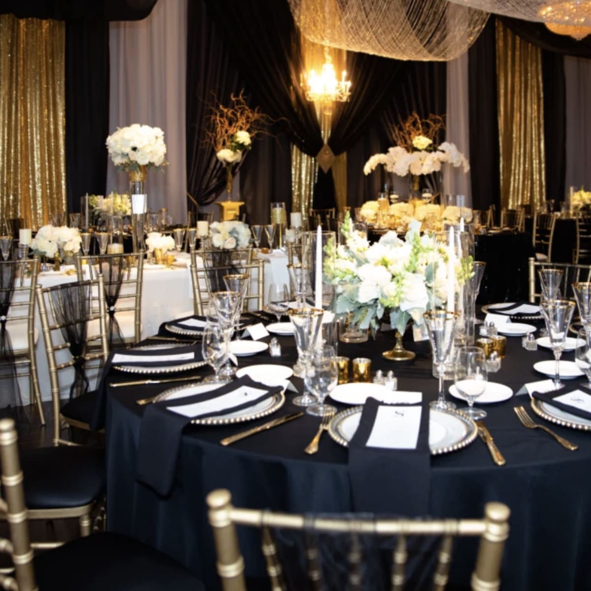 A banquet hall features elegantly arranged tables with black and gold decor, white floral centerpieces, and shimmering drapes.