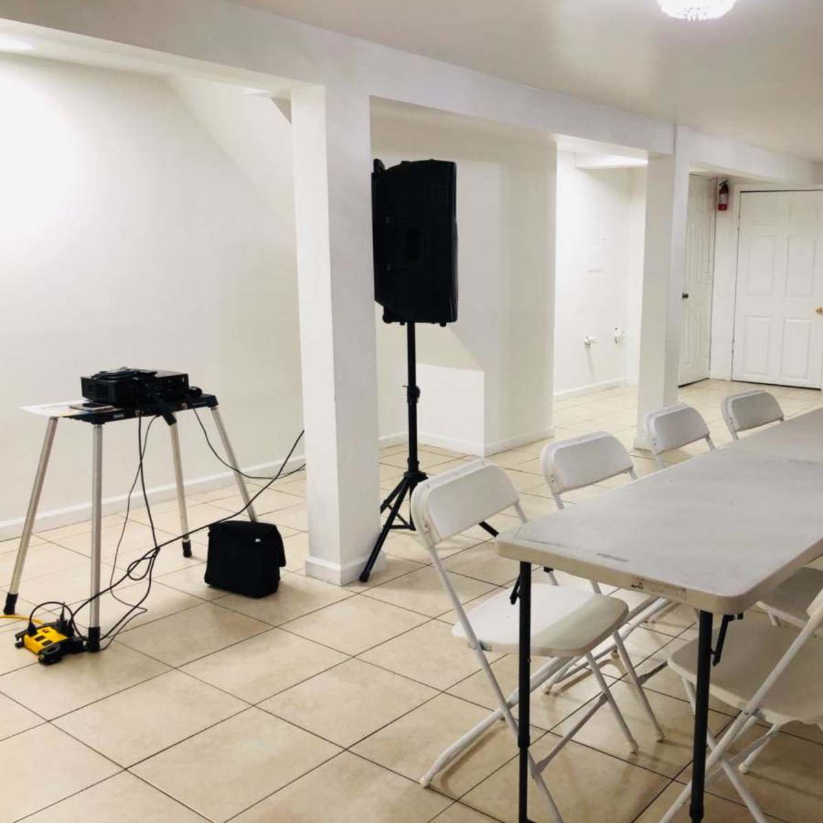 A row of white folding chairs is set around a long table in a well-lit, empty room with tiled flooring and a sound system on a nearby stand.