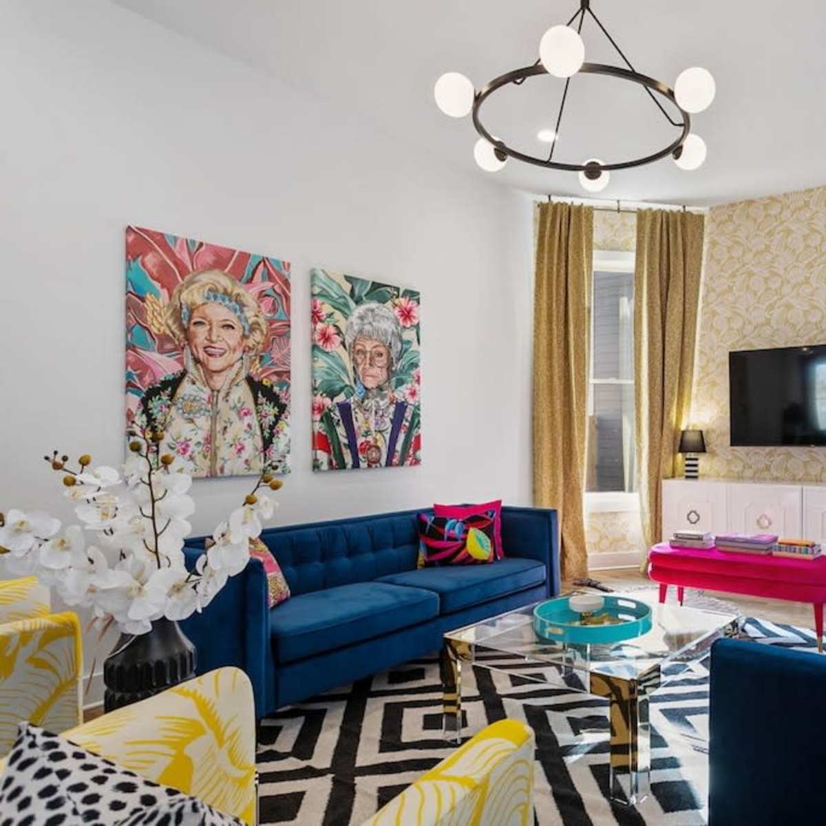 A brightly decorated living room featuring blue sofas, colorful cushions, patterned curtains, and wall art, with a television mounted above a white cabinet.
