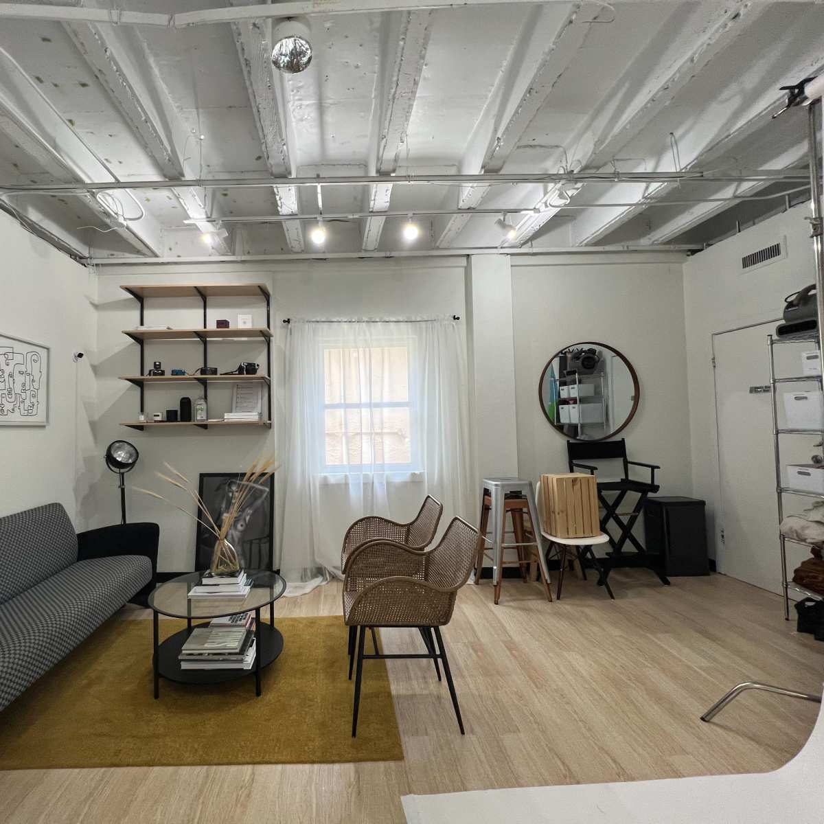 The room features a gray sofa, a round glass coffee table, and several chairs arranged around a window with sheer curtains, complemented by shelves and a mirror on the walls.