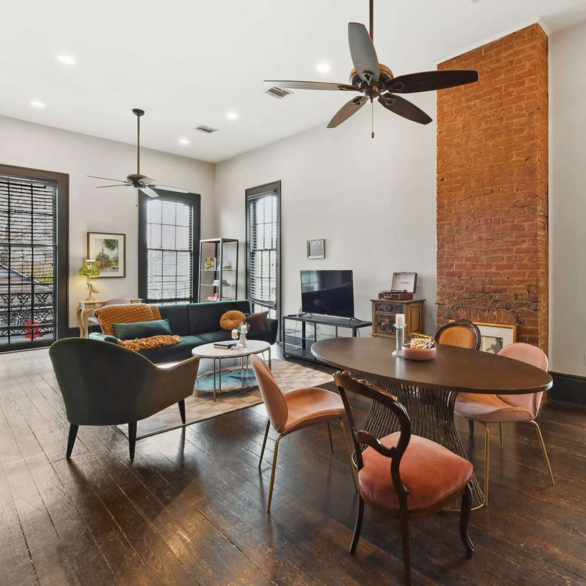 A spacious room features a dining table, chairs, a couch, and a television, with exposed brick walls and large windows adorned with shutters.