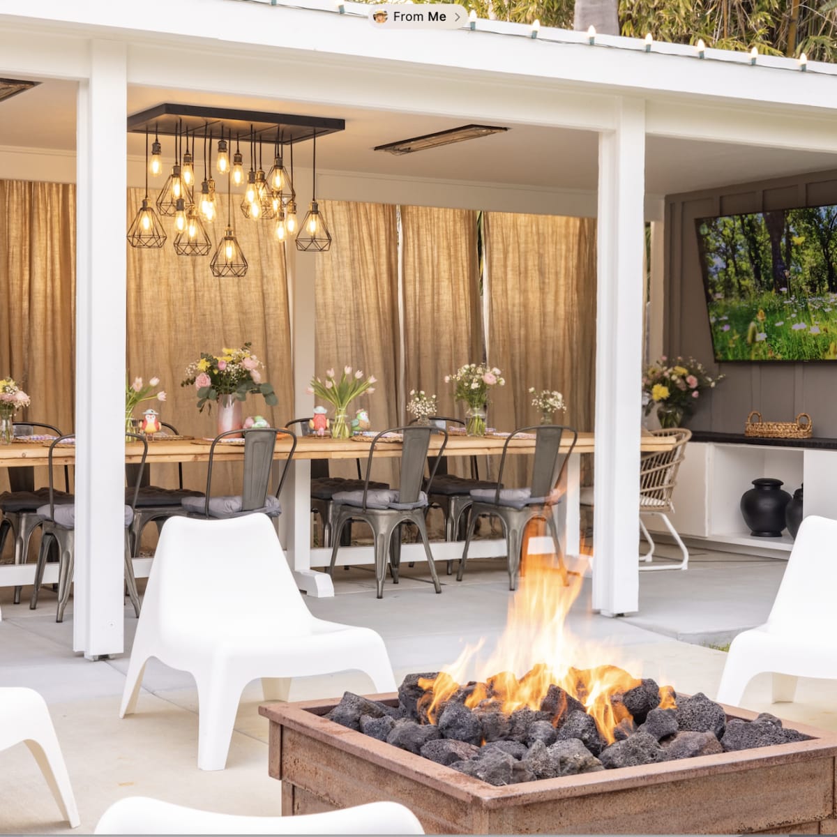 An outdoor dining area features a long wooden table with chairs, a fire pit with glowing coals, and a wall-mounted television displaying a landscape.