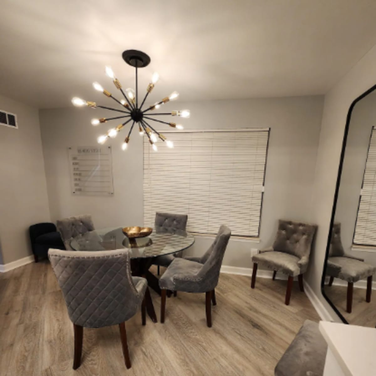 A modern dining area features a round glass table surrounded by six upholstered chairs, with a large chandelier and a mirror on the wall.