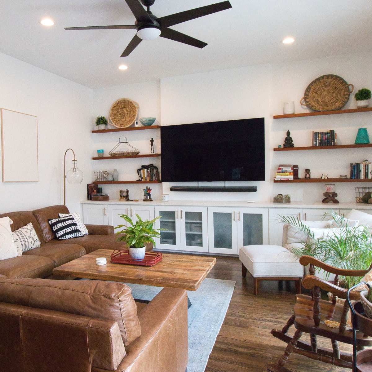 A modern living room features a brown leather sectional sofa, a wooden coffee table, a large wall-mounted TV, and built-in shelves with decorative items.