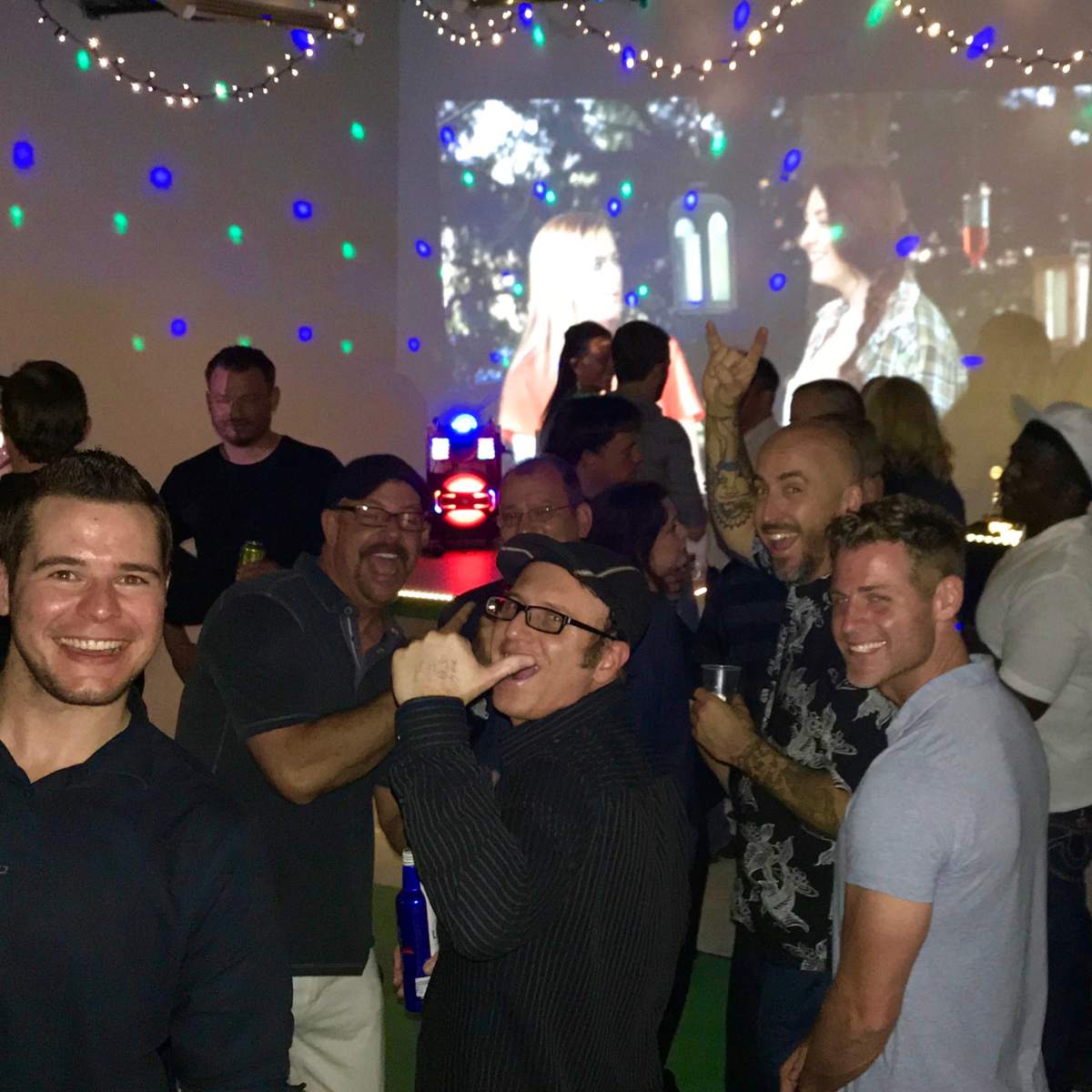 A group of people are socializing at a lively indoor party, with colorful lights and a movie projected on a wall behind them.