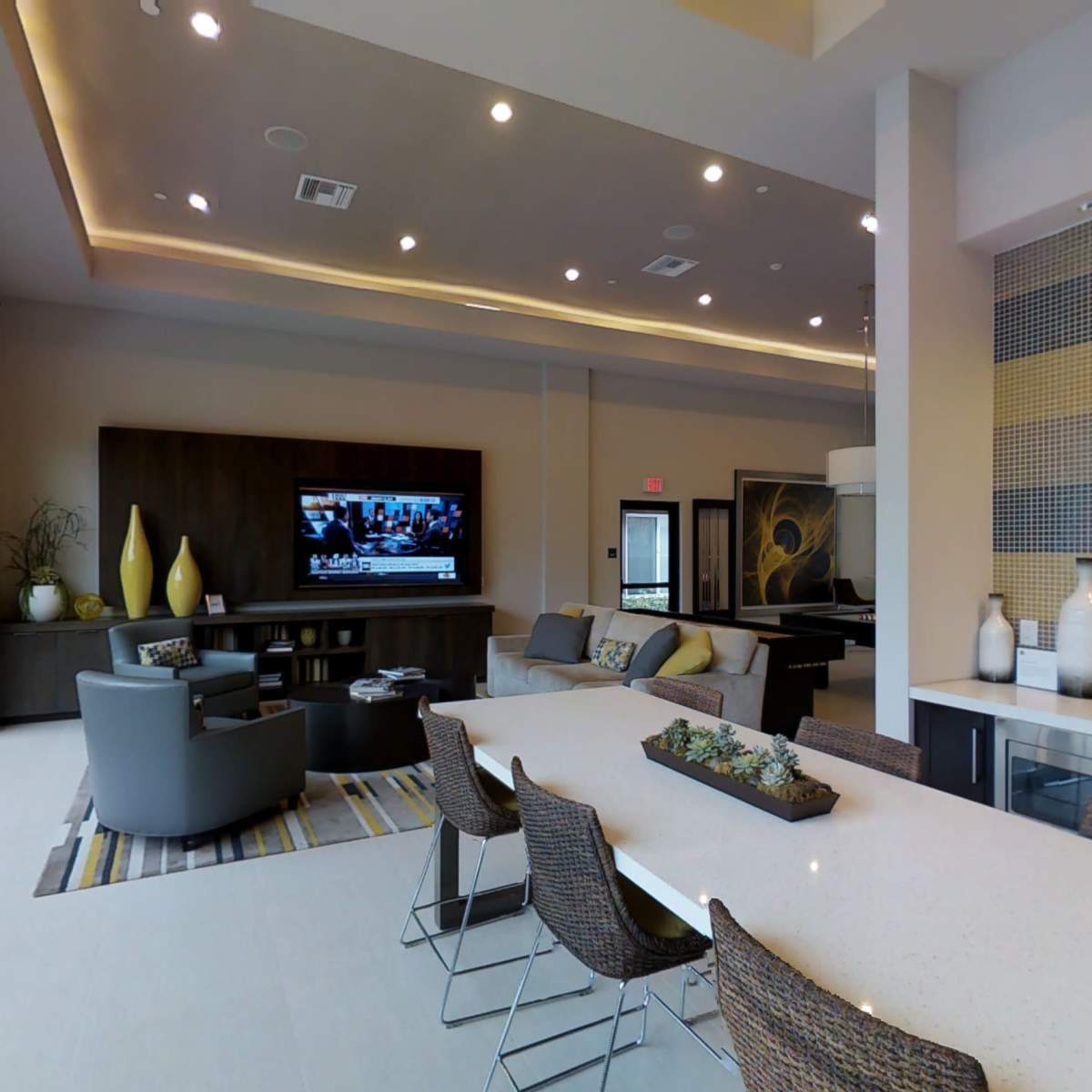 A modern lounge area with a large dining table, comfortable seating, a television on the wall, and large windows providing natural light.