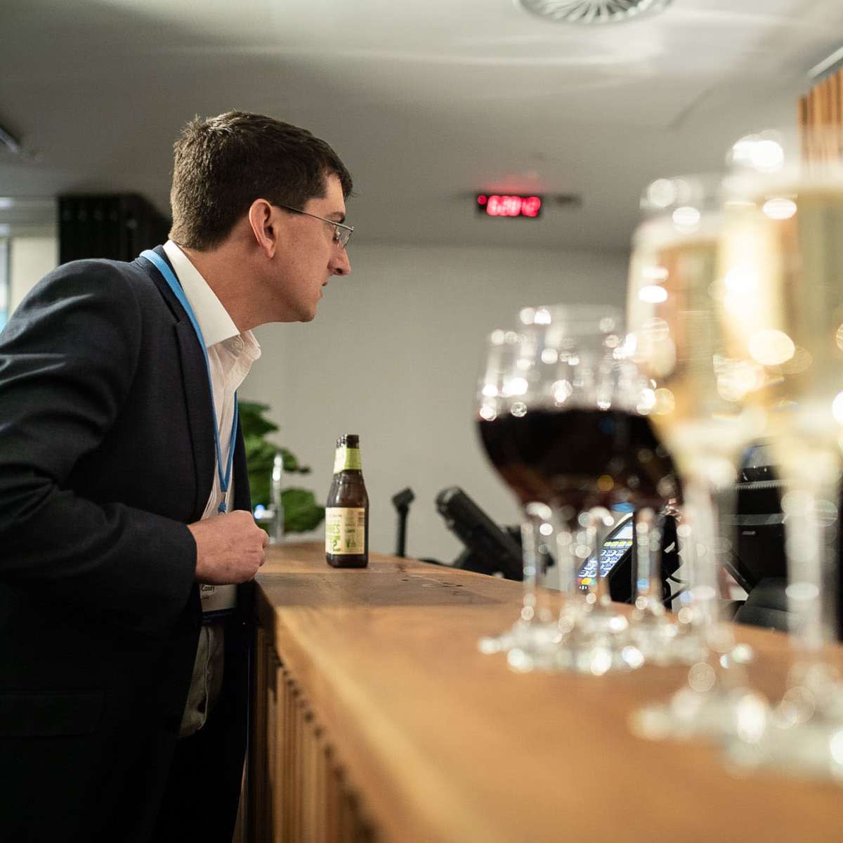 A man in a suit leans against a bar while observing the bartender prepare drinks, with various glasses of beverages lined up in the foreground.