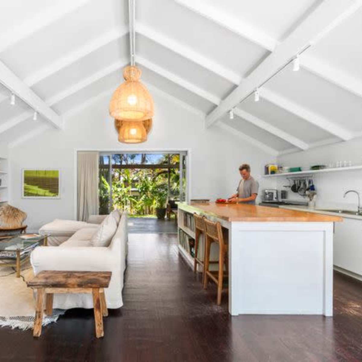 A modern living room features a white couch, a wooden coffee table, shelves filled with books, and a kitchen area with a counter where a person is preparing food.