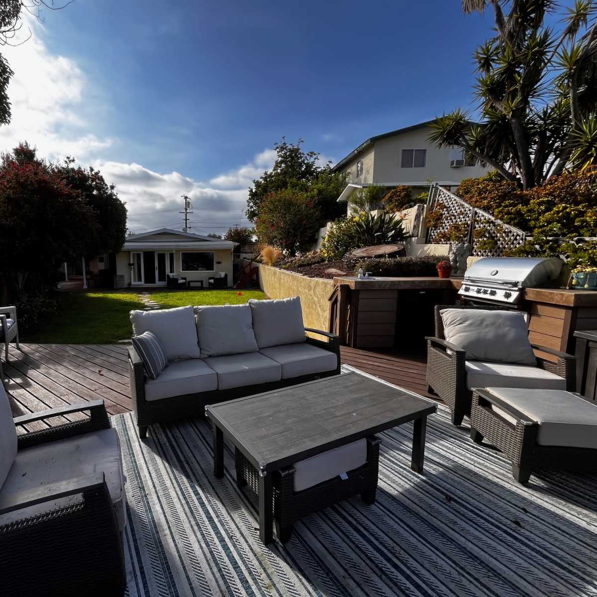 A cozy outdoor seating area with gray couches and a coffee table overlooks a well-maintained garden and a house in the background.