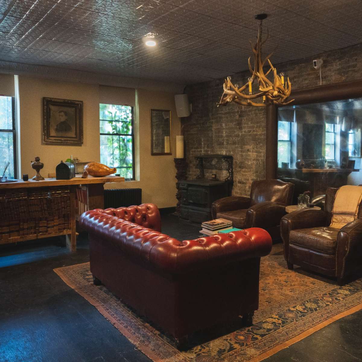 A cozy living room features a vintage leather sofa and armchairs, with a large mirror, a rustic chandelier, and exposed brick walls.