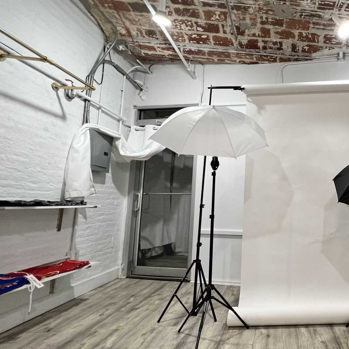 A photography studio with a white backdrop, two umbrellas (one white and one black) on tripods, and exposed brick walls.