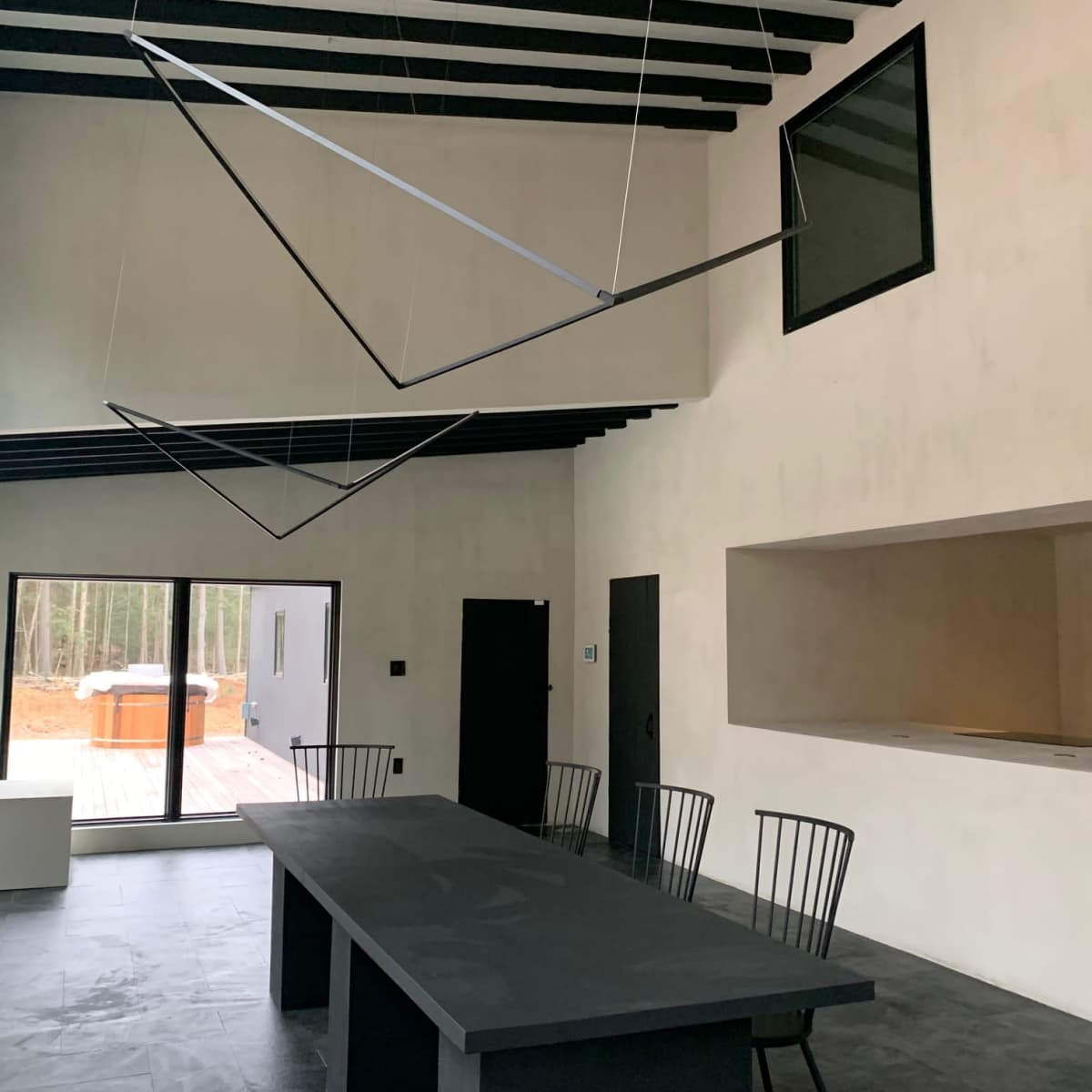 A modern interior space with high ceilings, featuring a long black table, four black chairs, and large windows that overlook a wooded area.