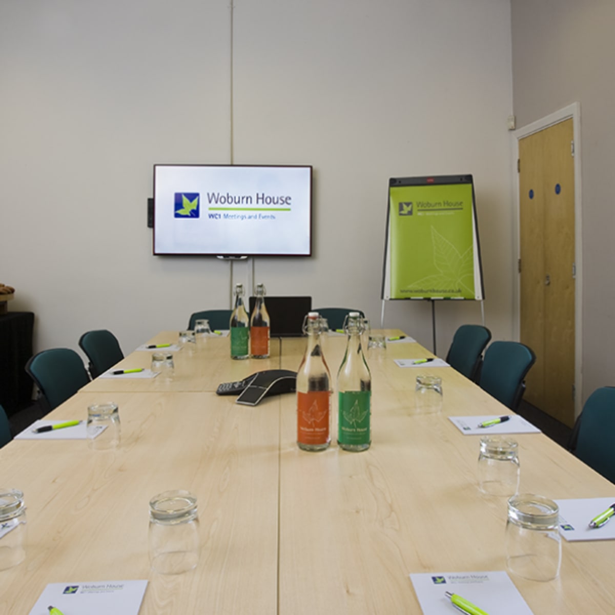 A long wooden conference table set with notebooks and water bottles, arranged in a meeting room featuring a large screen and a flip chart.