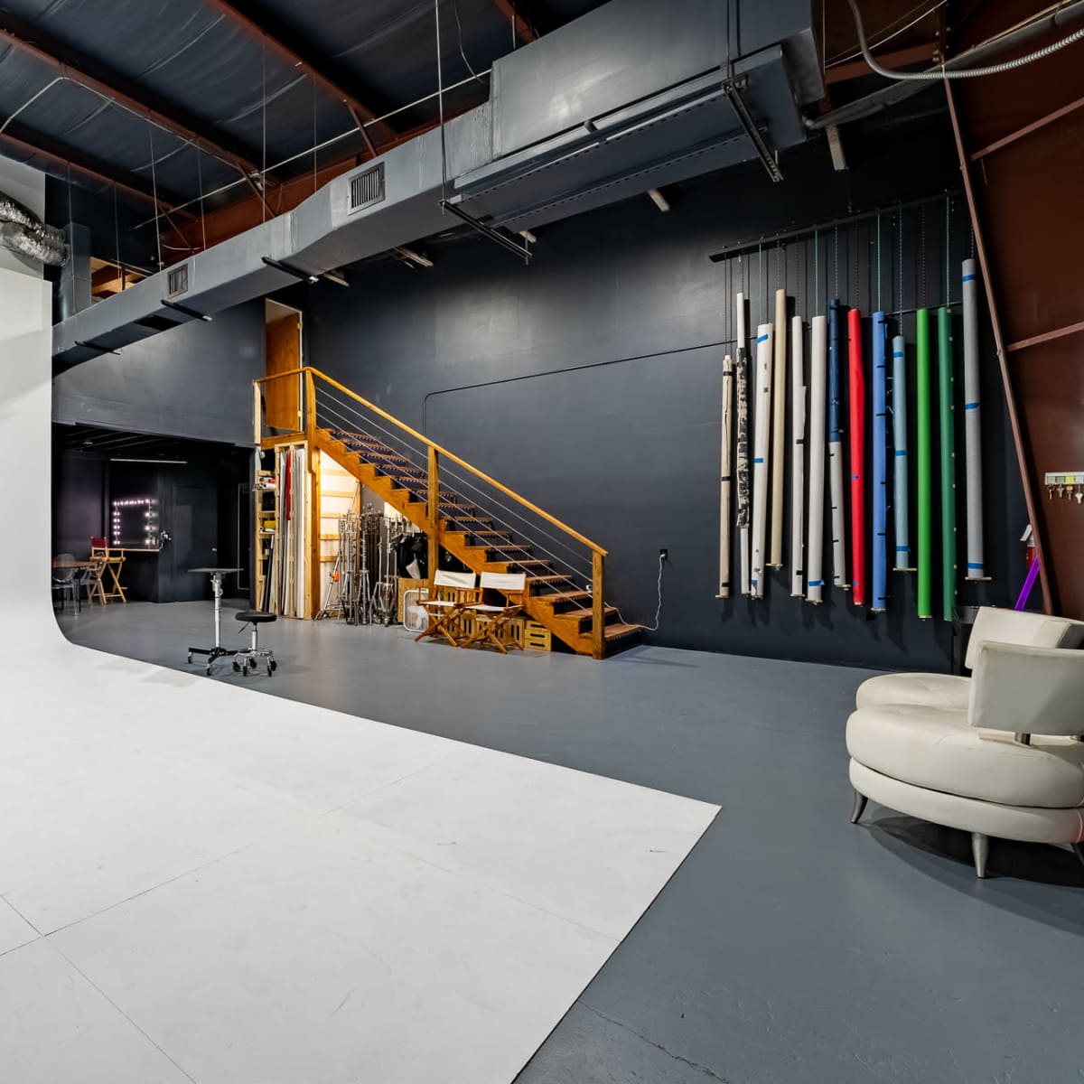 A photography studio with a large white backdrop, a wooden stool, a staircase leading to an upper level, and colorful rolls of backdrop material hanging on the wall.