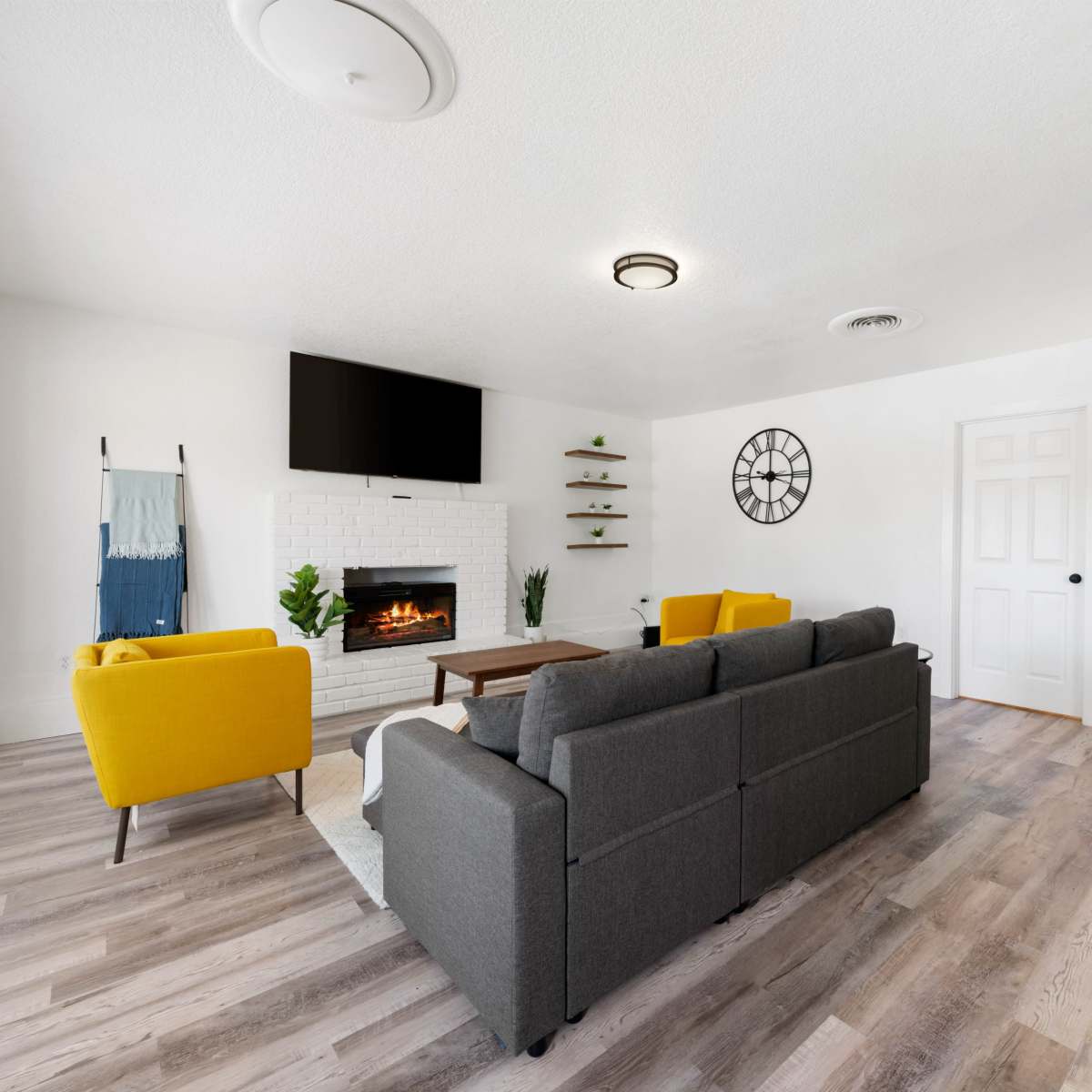 The living room features a modern design with a gray sofa, yellow chairs, a white brick fireplace, and a wall-mounted TV.