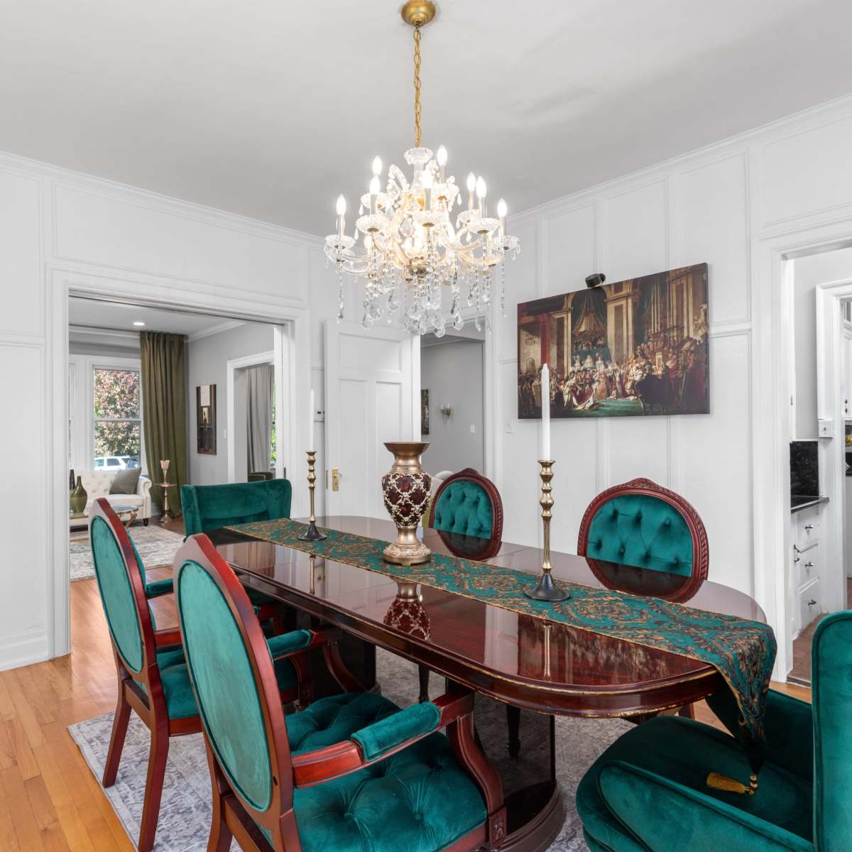 The dining room features a dark wooden table surrounded by six teal upholstered chairs and a crystal chandelier overhead.