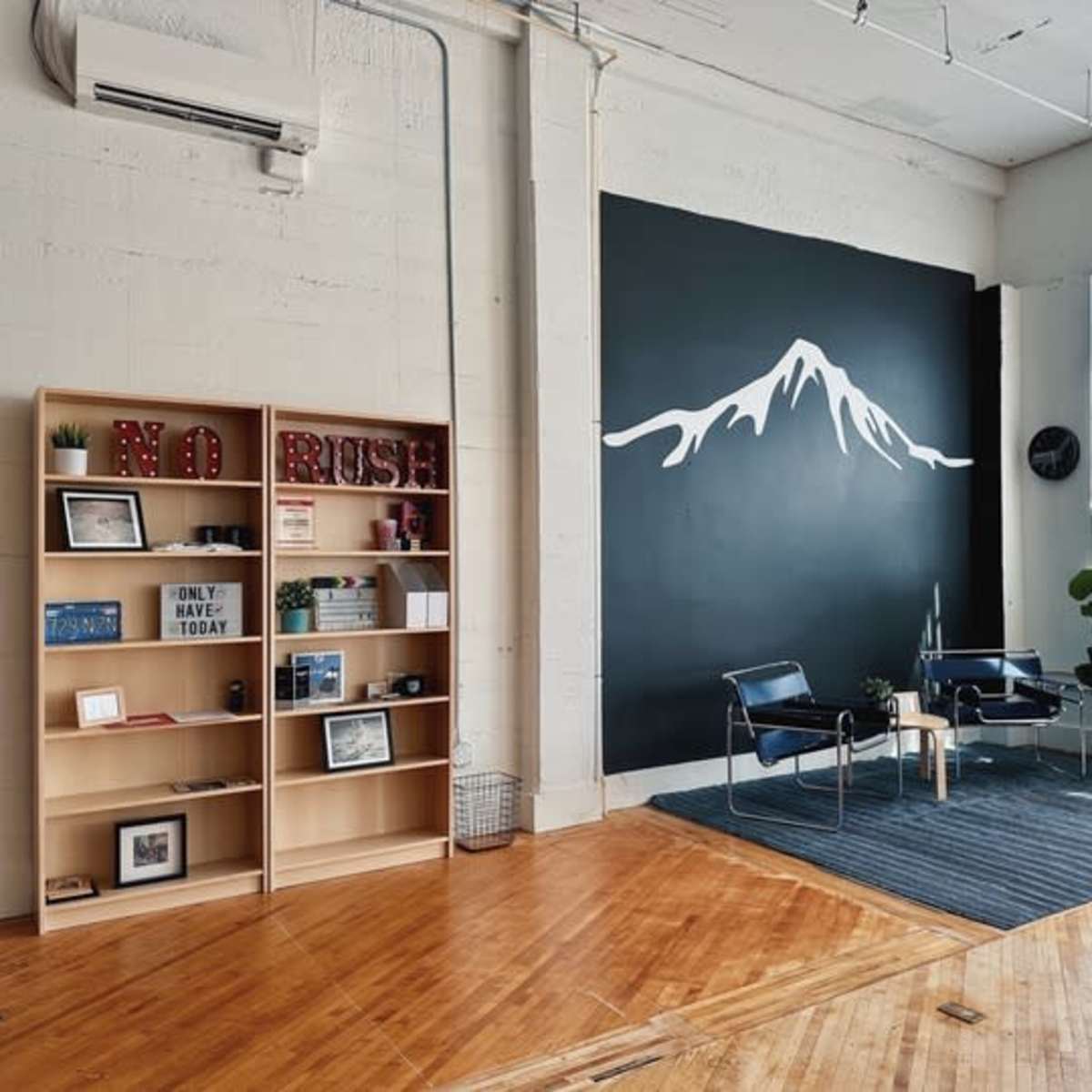 A minimalistic room with hardwood flooring, featuring a black wall painted with a mountain silhouette, a bookshelf filled with various items, and two chairs arranged around a small table on a striped rug.
