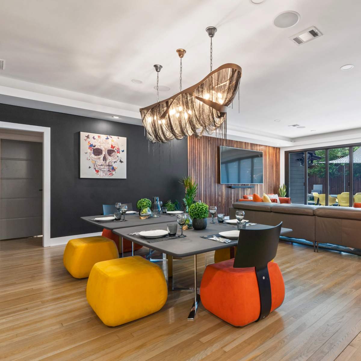 A modern living and dining area featuring a mix of colorful furniture, a large chandelier, and large glass doors leading to an outdoor space.