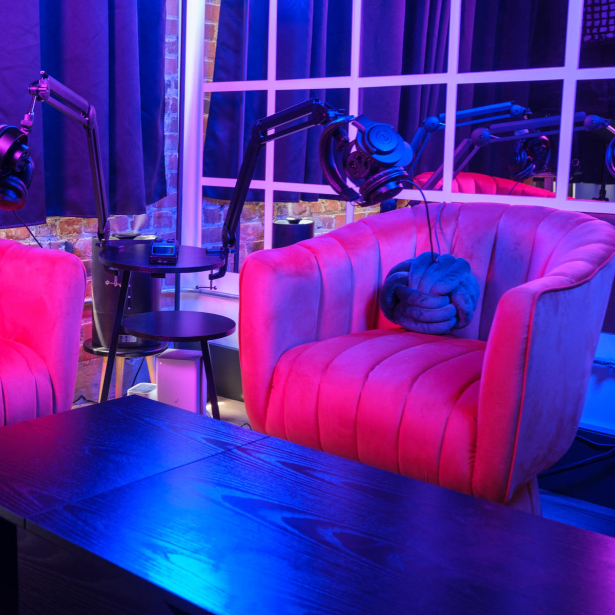 Two pink velvet chairs with microphones on adjustable arms are positioned around a black coffee table in a brightly lit room.