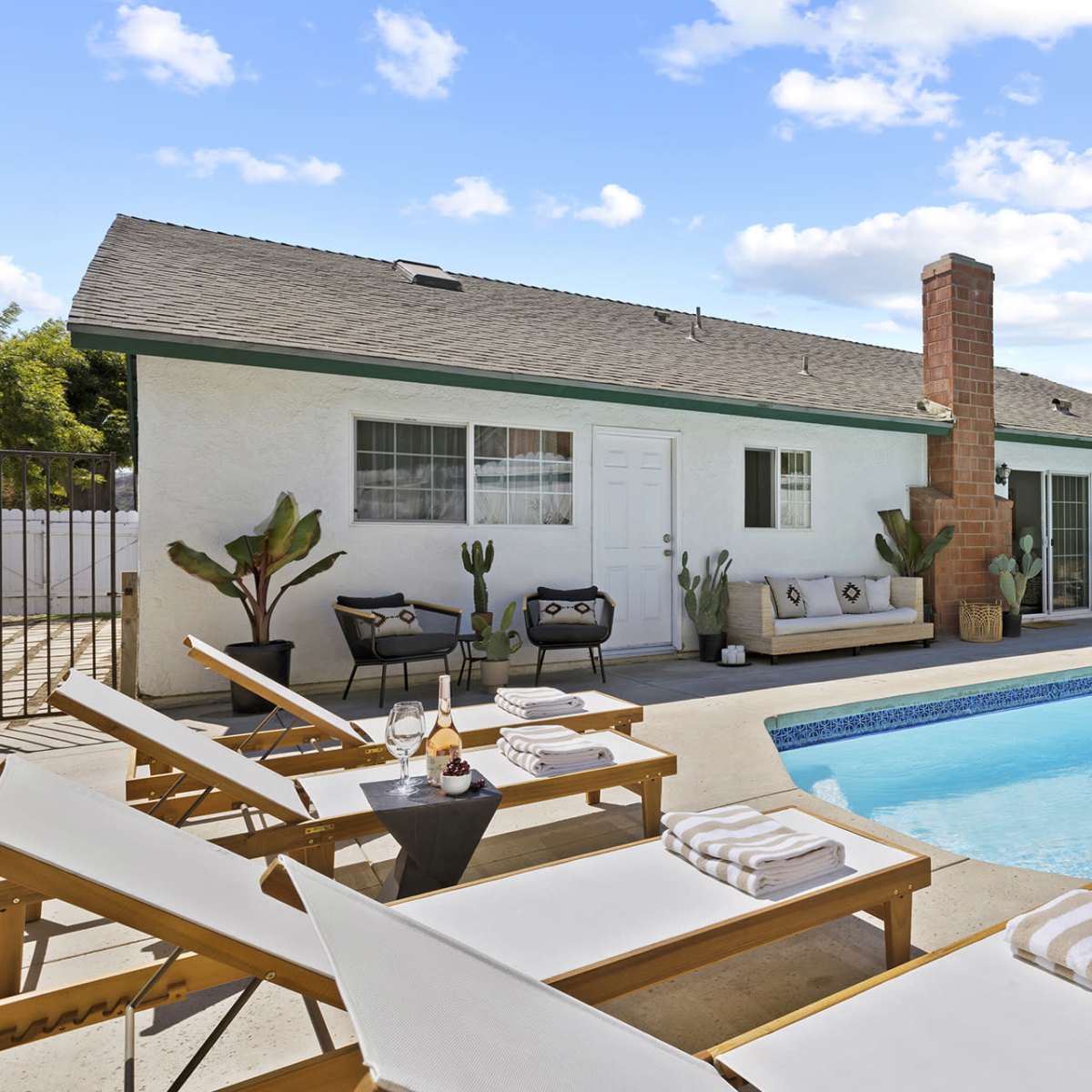 A backyard with a swimming pool, sun loungers, and a house with a chimney and potted plants.