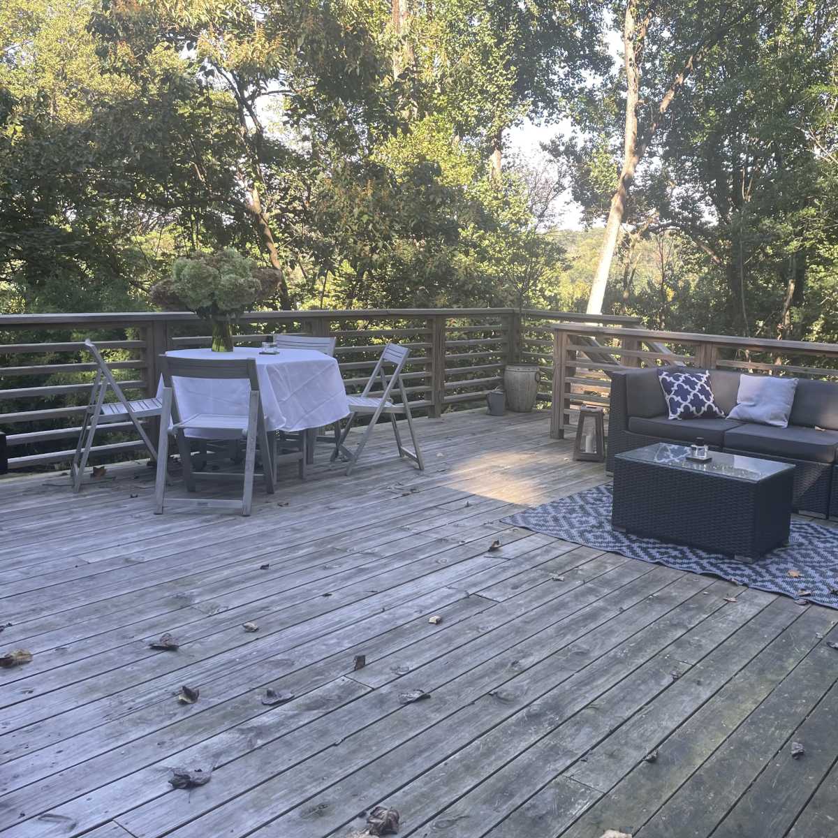 A wooden deck furnished with a sectional sofa, a coffee table, and dining setup, surrounded by trees.