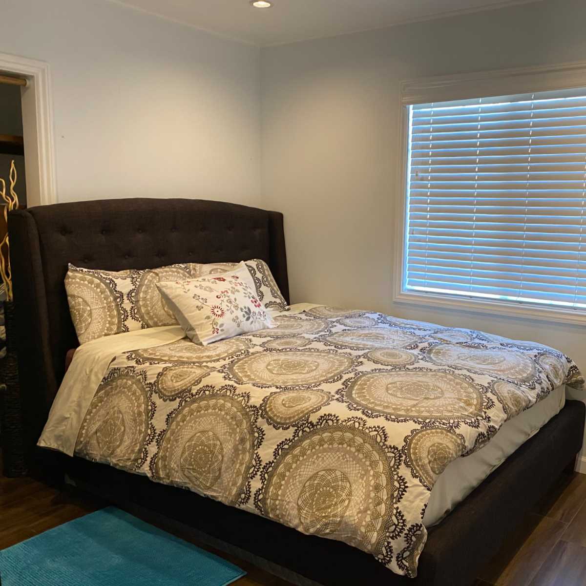 A neatly made bed with a patterned comforter is positioned against a wall in a well-lit bedroom, featuring a large mirror and a window with horizontal blinds.