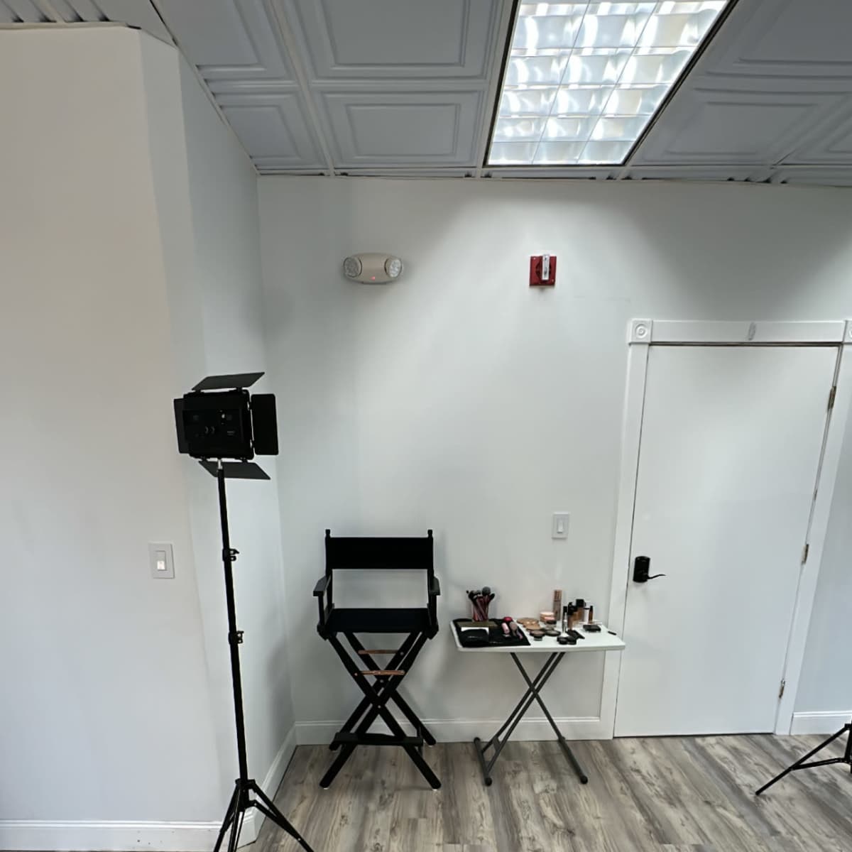 A minimalist makeup studio with a tall director's chair, a small table with makeup supplies, and two camera tripods on a wooden floor.